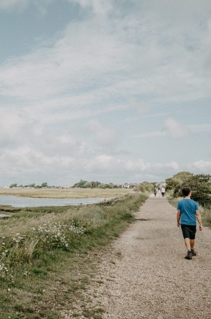 A man walking along a dusty path next to some grass and a stretch of beach, with boats in the distance.